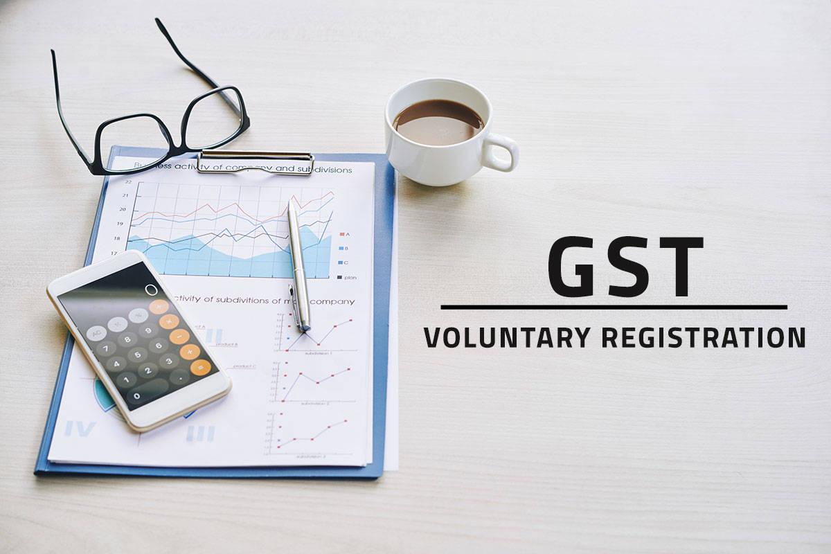 black text saying GST voluntary registration overlaying a white background with various items on a desk including black glasses, a filled coffee cup, a business financial report and a smartphone opened to a calculator app